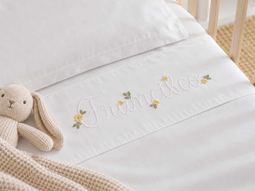 Lençol branco de berço com nome e pequenas flores amarelas bordadas junto à dobra