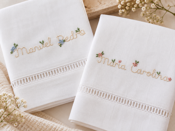 Duas toalhas brancas de batizado com nomes personalizados e pequenas flores bordadas